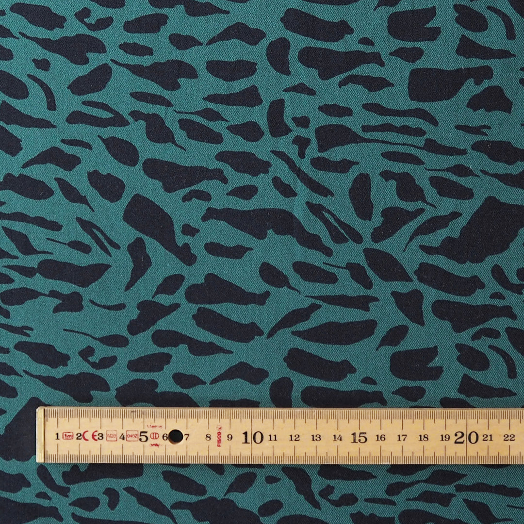 A close-up of fabric with an irregular dark abstract pattern on a teal background, with a wooden ruler along the bottom edge for scale.