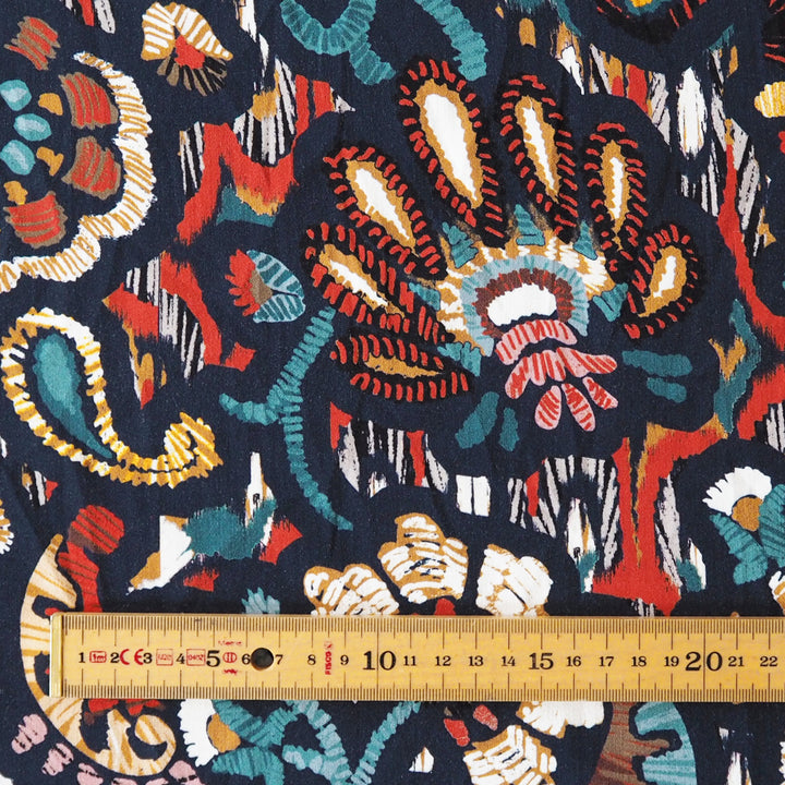 A close-up of a dark navy patterned fabric with colorful abstract paisley and floral motifs, shown with a wooden ruler at the bottom for scale.
