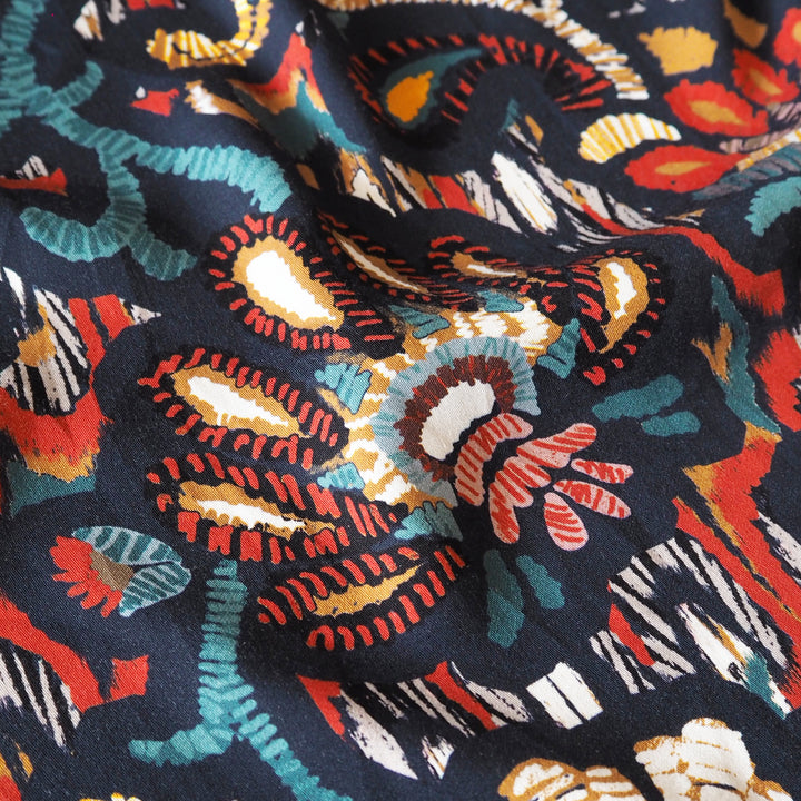 A close-up of a draped fabric with a dark navy blue base and a vibrant, multicolored ikat-style pattern featuring red, orange, teal, cream, and white motifs.