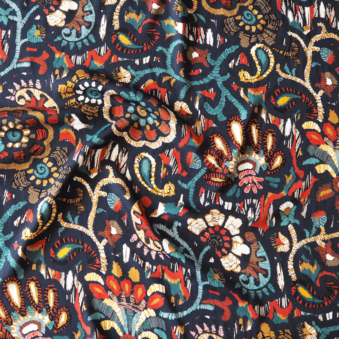 A close-up of draped fabric featuring a dense, hand-drawn style floral and paisley pattern in red, teal, cream, orange, and brown on a dark navy background.