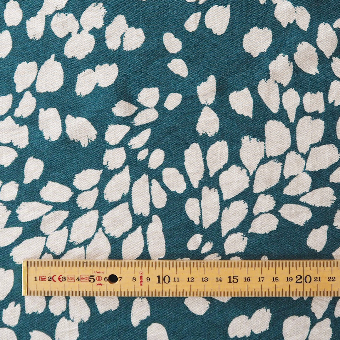 A close-up of teal fabric with an irregular white abstract leaf or petal pattern, with a wooden ruler along the bottom edge for scale.