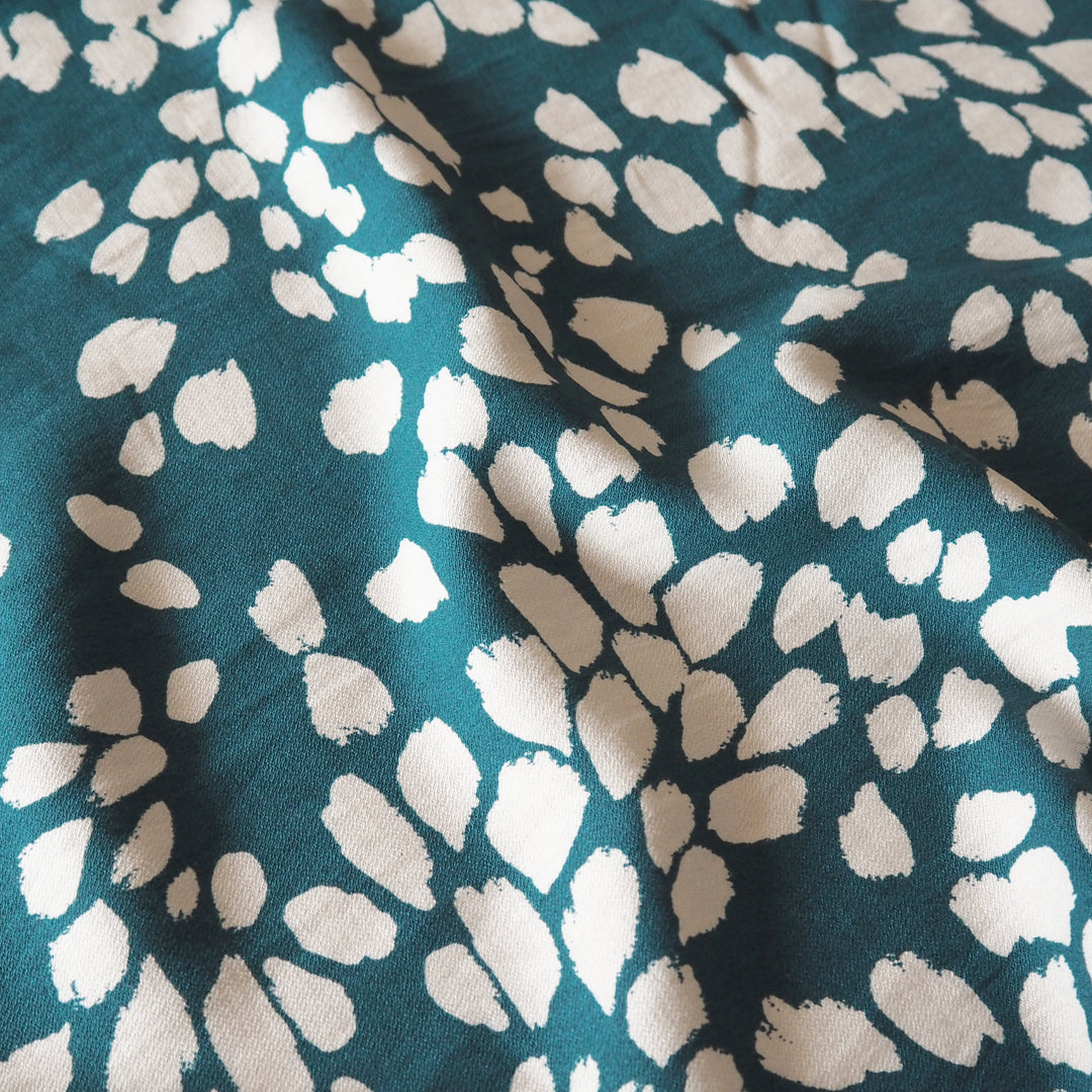 A close-up of a teal fabric with a white abstract spotted pattern, showing soft folds and texture.