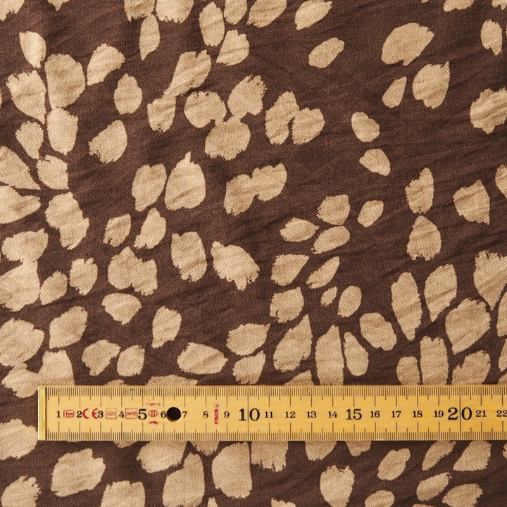 Brown fabric with a beige irregular spotted animal print pattern, shown with a yellow measuring tape ruler at the bottom for scale.