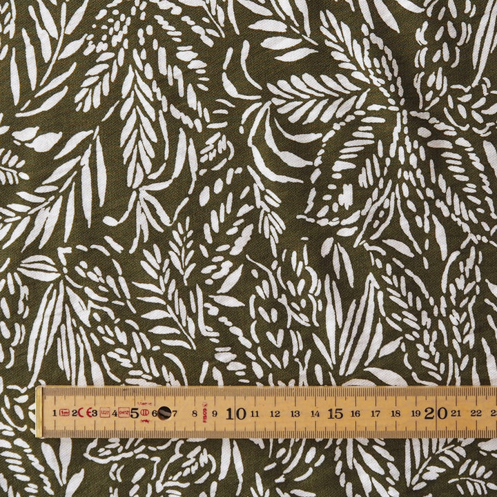 A close-up of dark olive green fabric with a white leafy botanical pattern, shown with a wooden ruler along the bottom edge for scale.