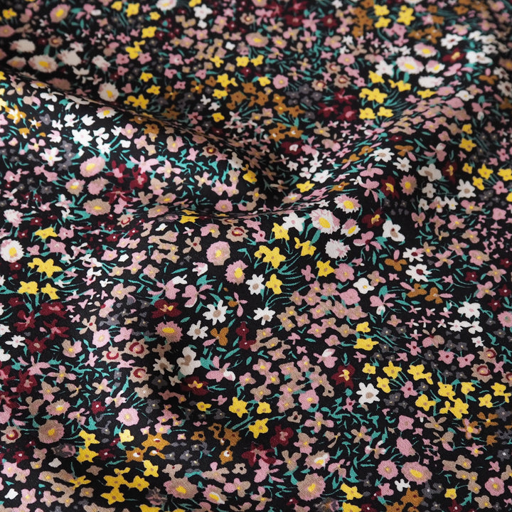 A close-up of a black fabric with a dense, multicolor floral print featuring pink, yellow, white, and burgundy flowers with green stems.