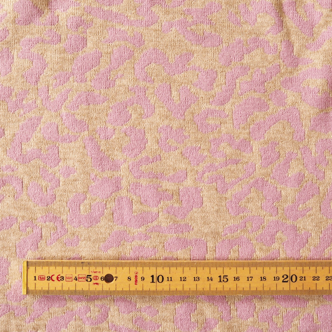 A close-up of woven fabric with an abstract leopard-like pattern in soft pink and warm beige, with a wooden tape measure along the bottom edge for scale.