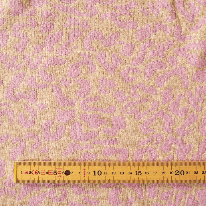 A close-up of woven fabric with an abstract leopard-like pattern in soft pink and warm beige, with a wooden tape measure along the bottom edge for scale.