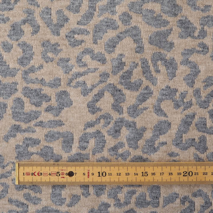 A close-up of a woven fabric with an abstract blue-gray and beige pattern, shown with a wooden ruler along the bottom edge for scale.