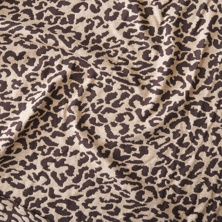A close-up of leopard print fabric with dark brown irregular spots on a warm beige background, shown with soft folds and texture.