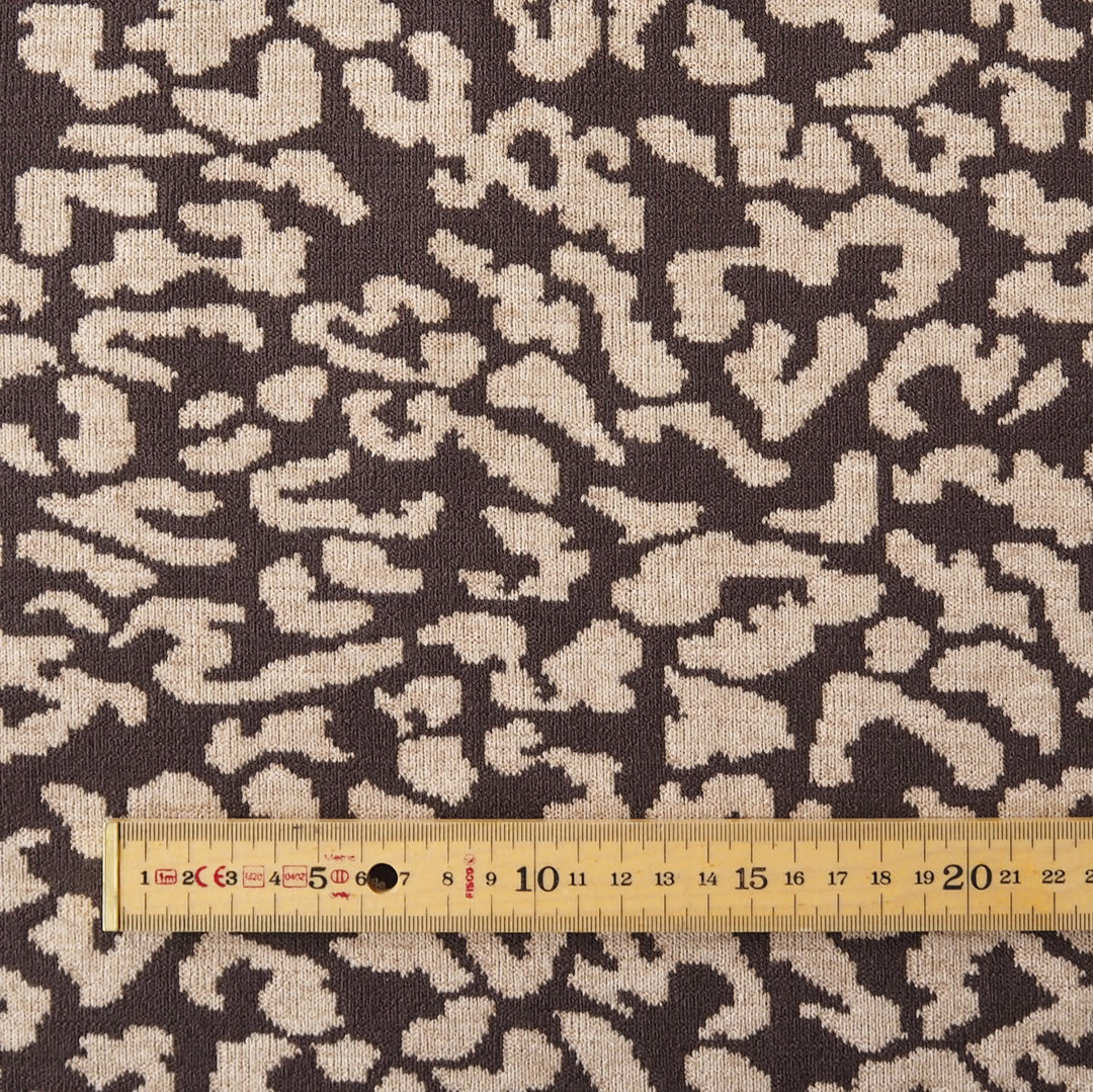 Close-up of a woven fabric with an abstract organic pattern in dark brown and warm beige, with a wooden ruler along the bottom edge for scale.