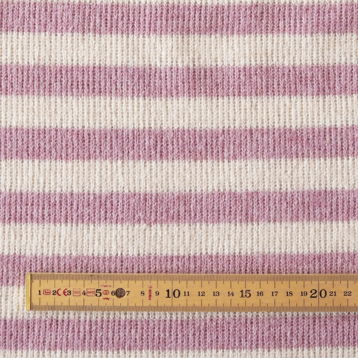 Close-up of a knitted fabric with horizontal stripes in dusty pink and off-white, with a wooden ruler at the bottom showing the scale.