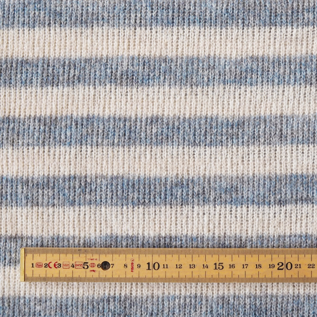Close-up of a striped knit fabric with horizontal bands of light blue, medium blue, and off-white, shown with a wooden ruler at the bottom for scale.