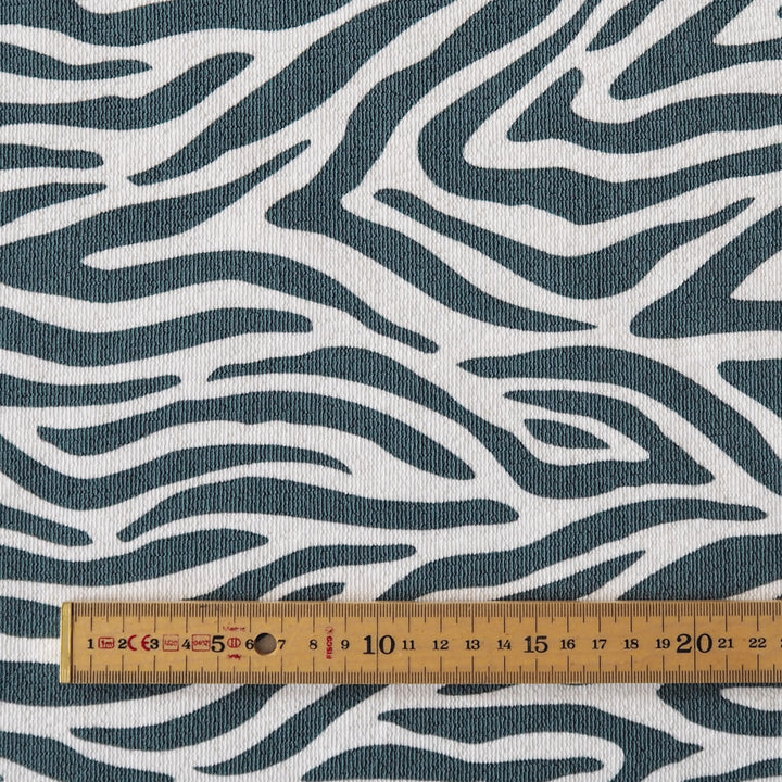 A close-up of a fabric with a wavy zebra-like pattern in dark teal and off-white, with a wooden ruler at the bottom for scale.