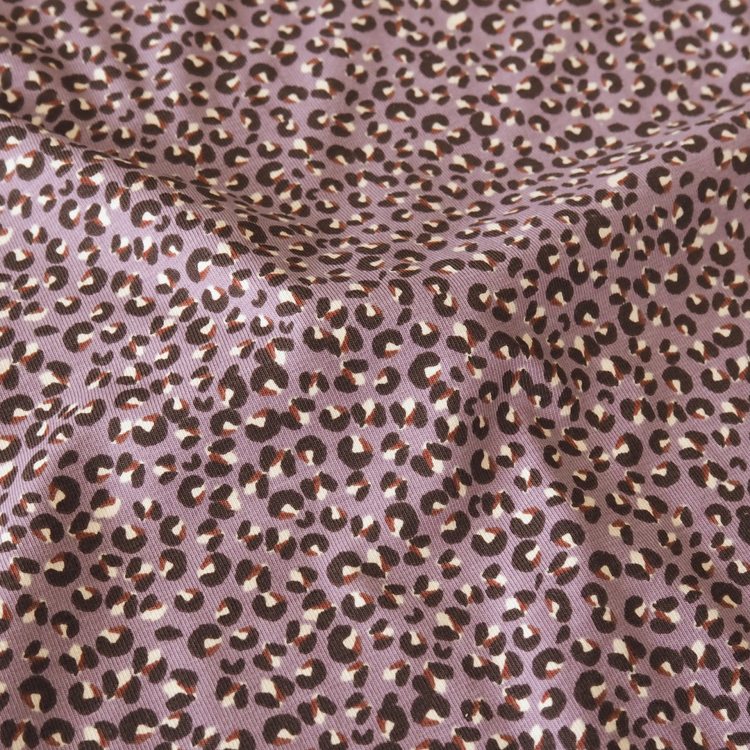 A close-up of leopard-print fabric with a dusty mauve-purple base, dark brown spots, and lighter cream highlights, shown with soft folds.