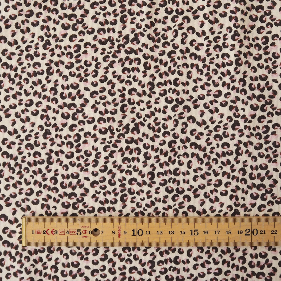 A close-up of leopard-print fabric with a cream background and dark brown to black spots, shown with a wooden ruler along the bottom edge for scale.