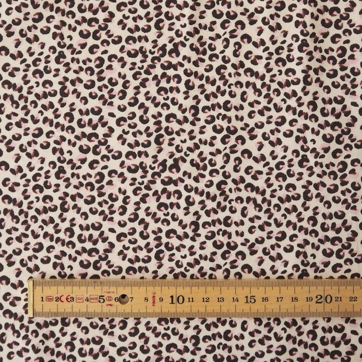 A close-up of leopard-print fabric with a cream background and dark brown to black spots, shown with a wooden ruler along the bottom edge for scale.