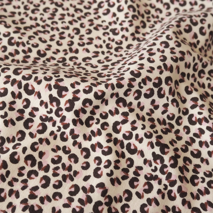 A close-up of leopard print fabric with dark brown spots on a warm beige background, featuring soft folds and a subtle pinkish cast in some areas.
