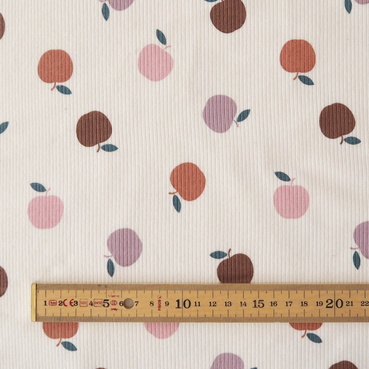 A close-up of a ribbed fabric with a repeating pattern of stylized fruit in pink, brown, and purple on an off-white background, with a wooden ruler at the bottom for scale.
