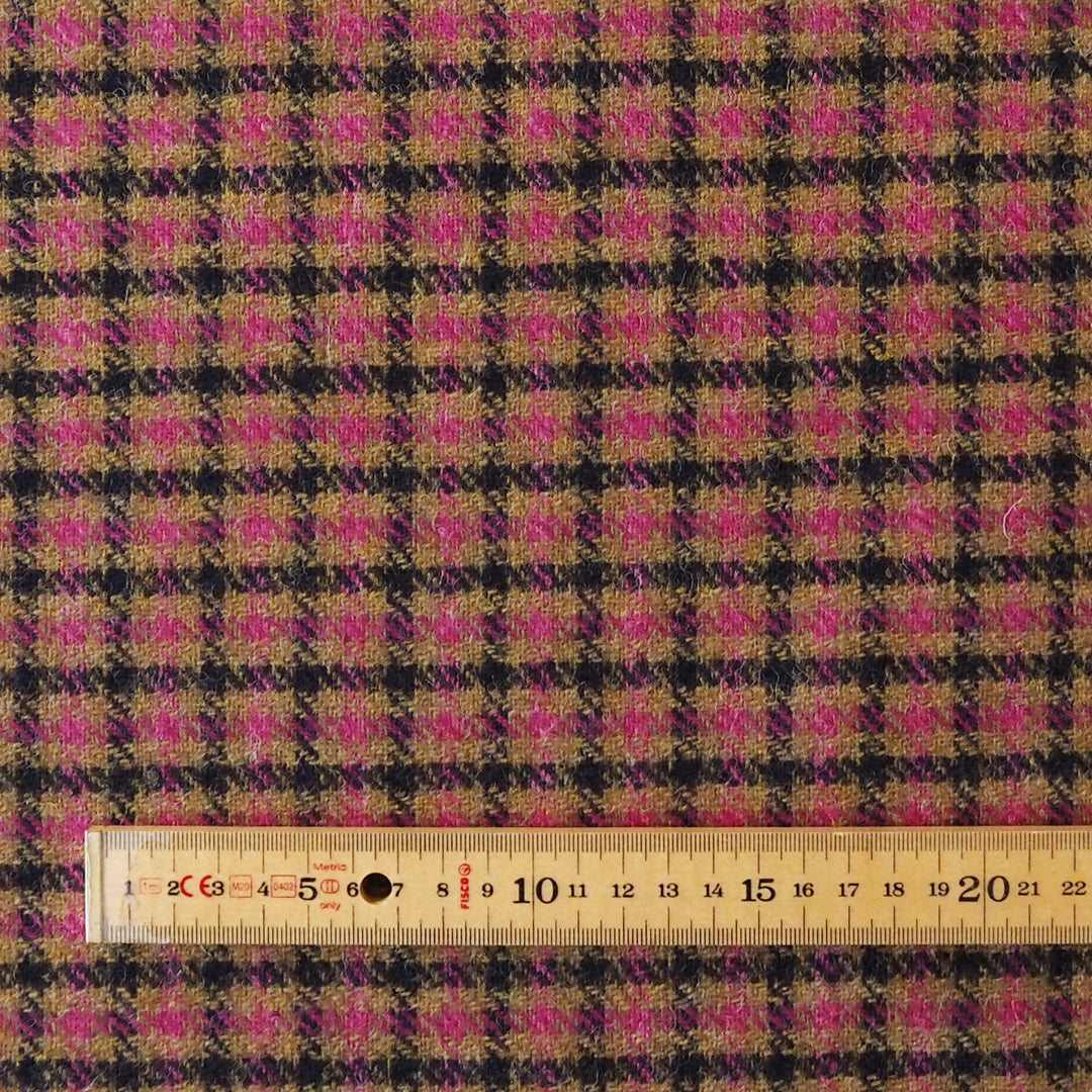 A close-up of a plaid fabric with a repeating check pattern in black, tan, and bright pink, with a wooden ruler laid across the bottom edge for scale.