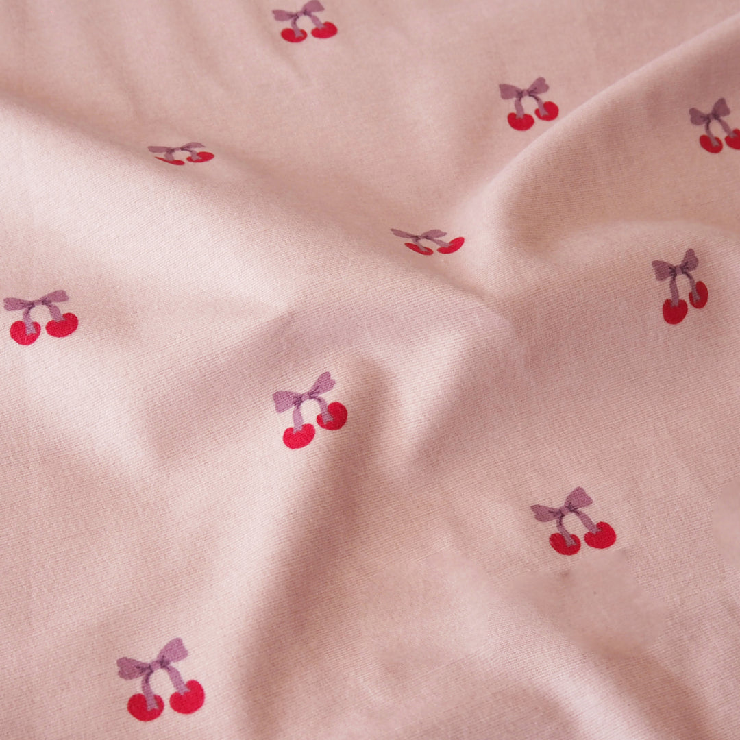 A close-up of a light pink fabric with a repeating pattern of small red cherries tied with purple bows, shown with soft folds.