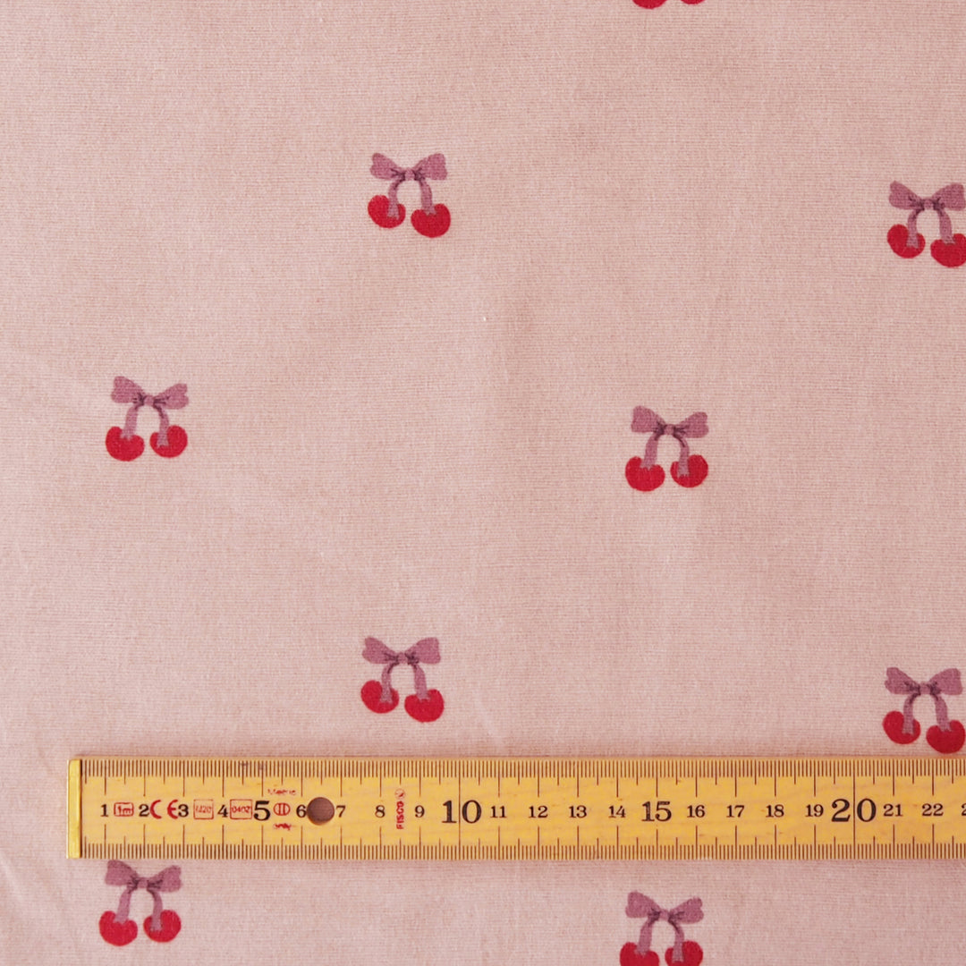 Pink fabric with a repeating cherry print, shown with a wooden ruler at the bottom to indicate scale.