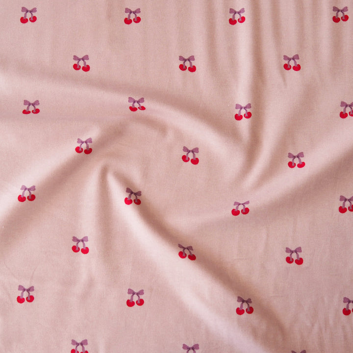 Light pink fabric with a repeating pattern of small red cherries tied with purple bows, shown with soft folds.