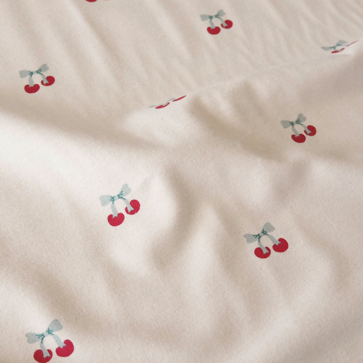 A close-up of a cream-colored fabric with a repeating pattern of small red cherries tied with light blue bows.