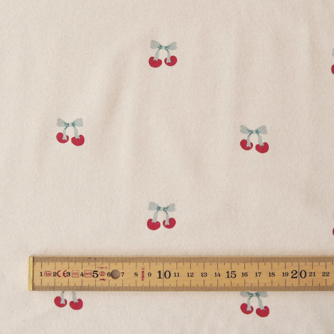 Beige fabric with a repeating pattern of small red cherries tied with light blue bows, shown with a wooden ruler at the bottom for scale.