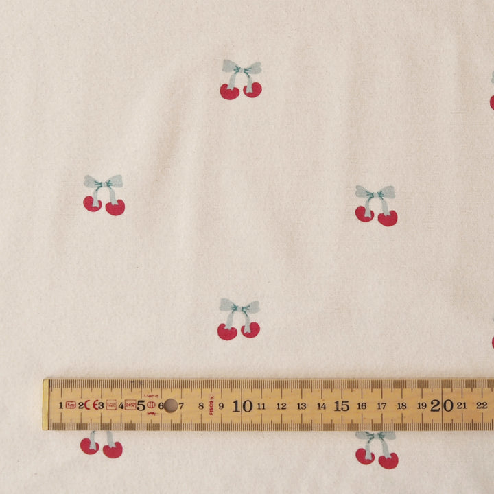 Beige fabric with a repeating pattern of small red cherries tied with light blue bows, shown with a wooden ruler at the bottom for scale.