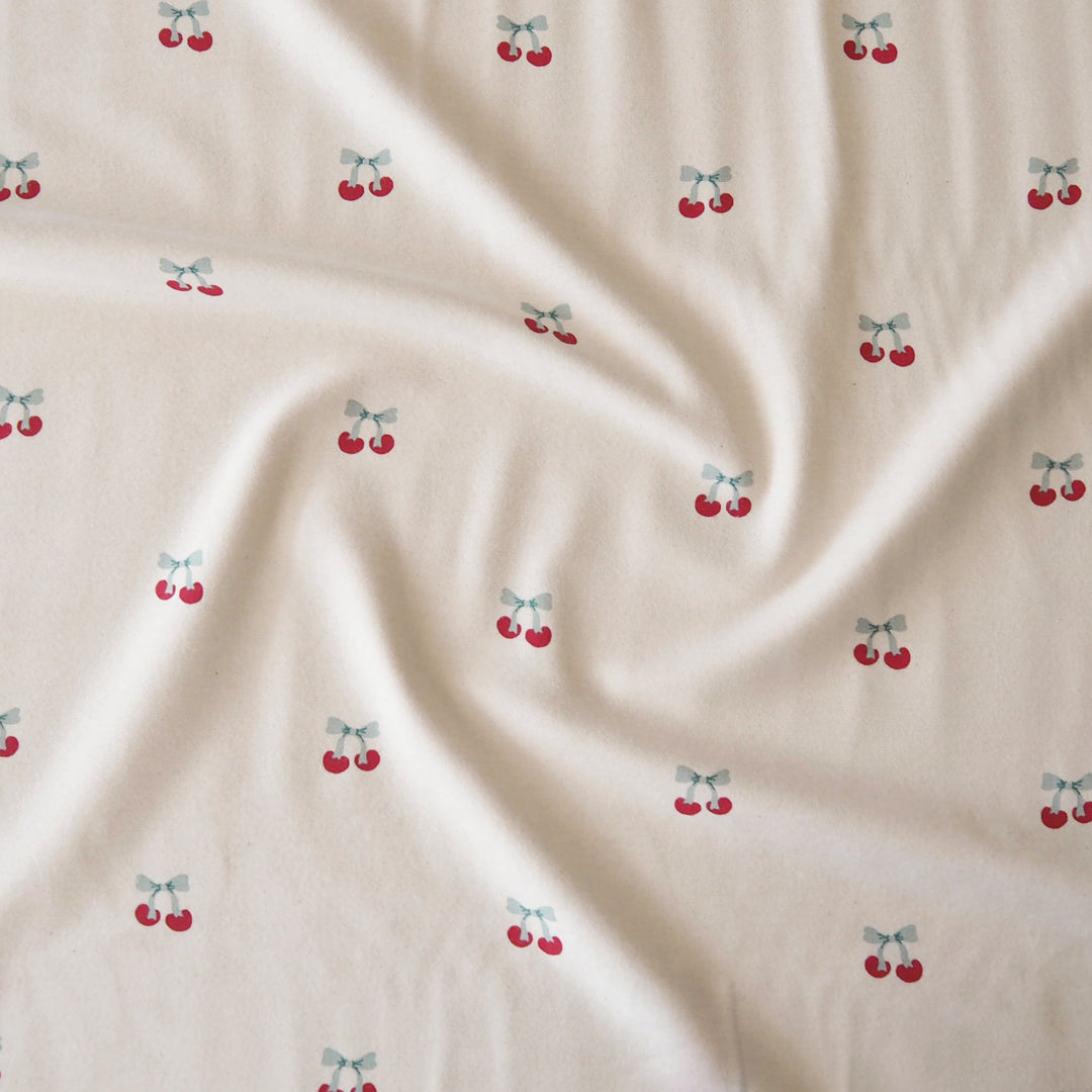 A close-up of a cream-colored fabric with a repeating pattern of small red cherries tied with light blue bows, shown with soft folds.