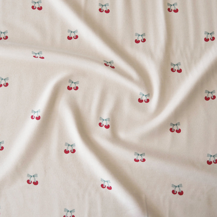 A close-up of a cream-colored fabric with a repeating pattern of small red cherries tied with light blue bows, shown with soft folds.