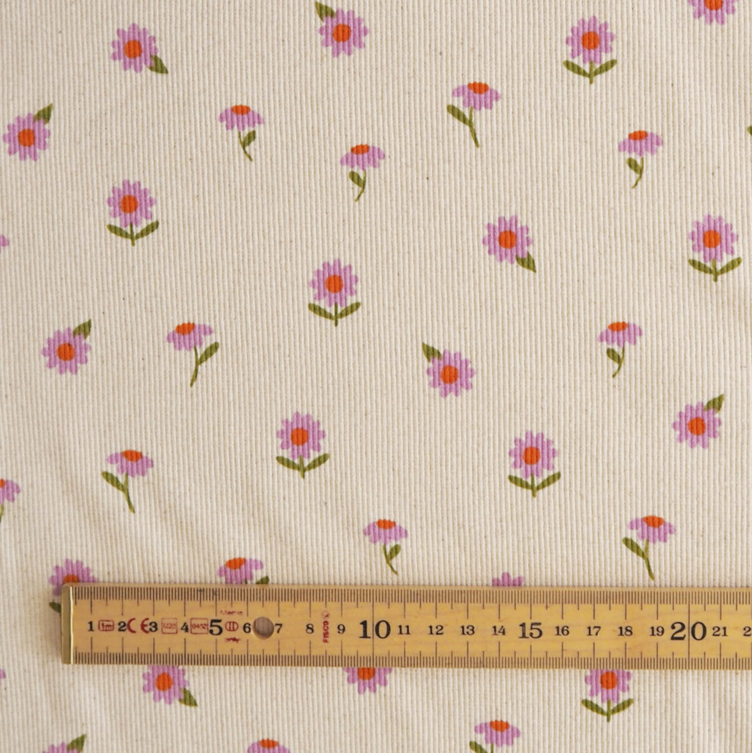 A close-up of cream-colored fabric with a repeating pattern of small pink and purple flowers with green stems, with a wooden ruler at the bottom for scale.