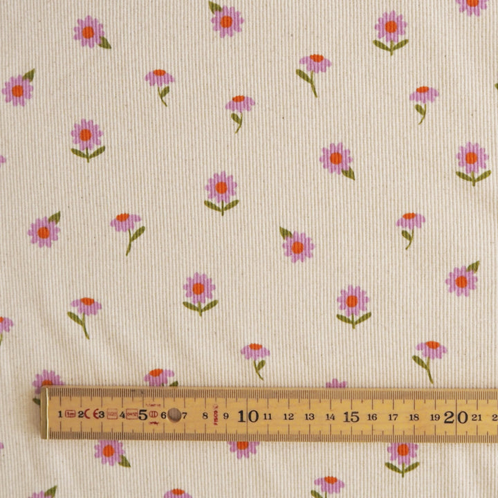 A close-up of cream-colored fabric with a repeating pattern of small pink and purple flowers with green stems, with a wooden ruler at the bottom for scale.