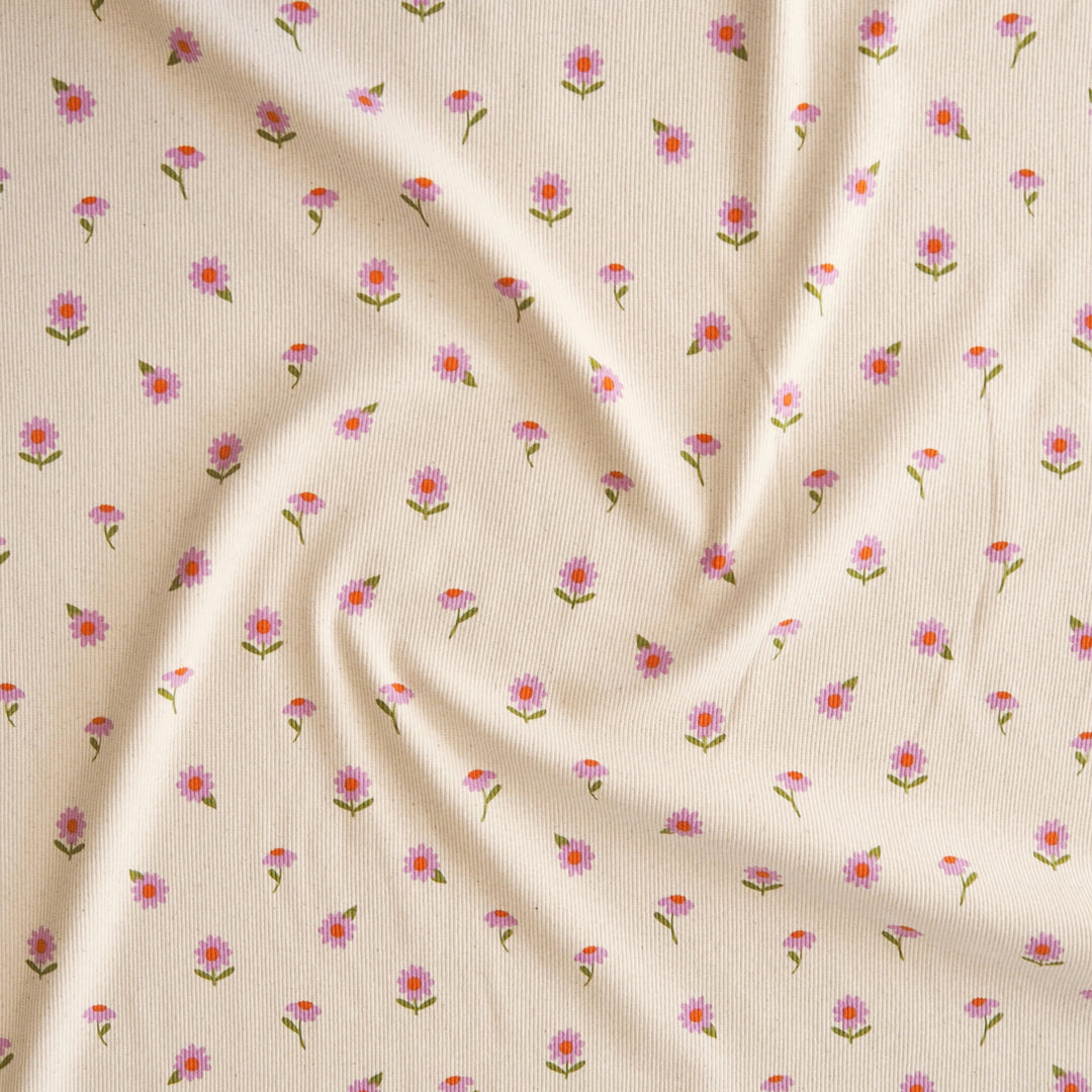 A close-up of a cream-colored fabric with a delicate, repeating pattern of small pink and purple flowers with green stems, arranged in soft folds.