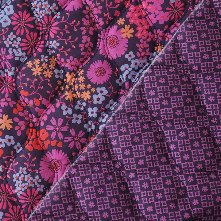 Close-up of a quilted fabric with a diagonal split design: one side shows a pink, purple, orange, and blue floral print on a dark background, and the other side shows a purple geometric lattice pattern on a dark background.