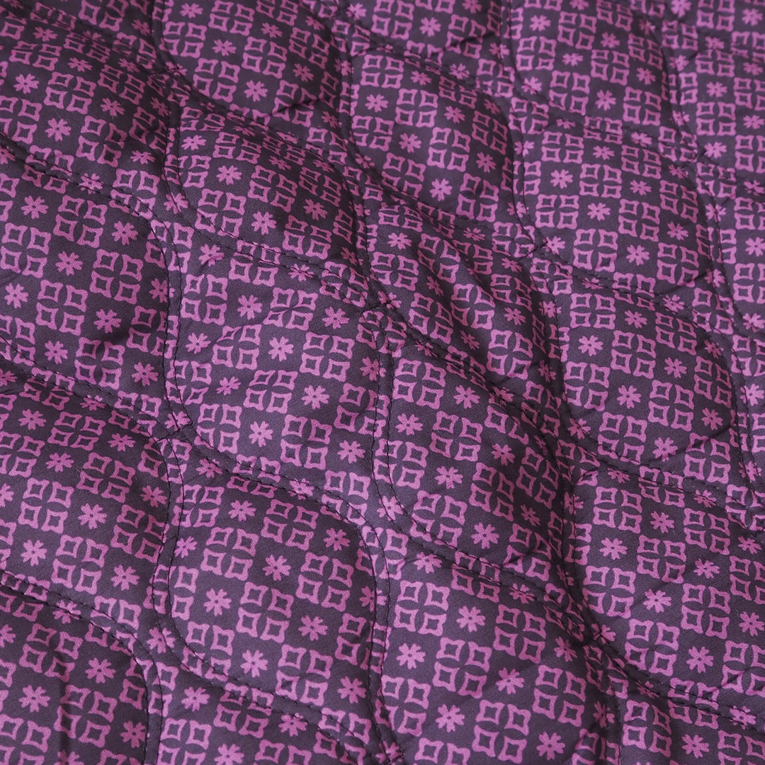 A close-up of a quilted fabric with a repeating geometric pattern in shades of purple and pink.