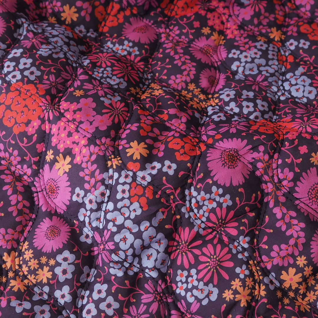 A close-up of a quilted fabric surface with a dense floral print in pink, orange, lavender, and white on a dark charcoal background.