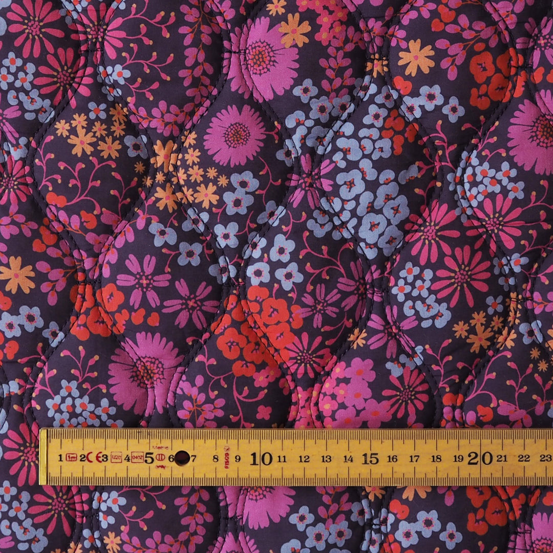 A close-up of a quilted fabric with a retro floral pattern in pink, orange, light blue, and yellow on a dark purple background, with a yellow measuring tape ruler placed along the bottom edge for scale.
