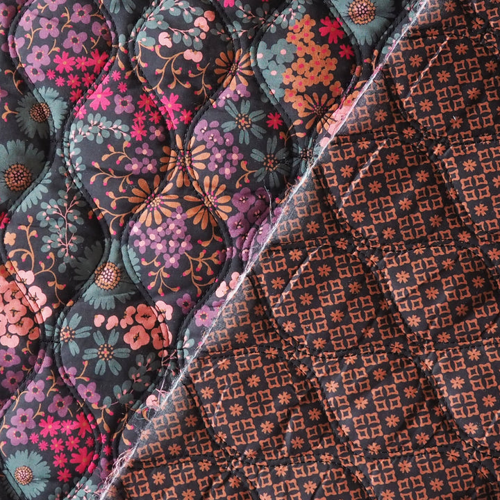 Close-up of a quilted fabric showing two patterned sides: one side has a floral print in pink, purple, teal, and orange on a dark background, and the other side has a repeating geometric floral pattern in orange on a dark background.