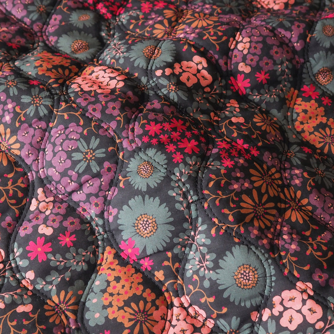 A close-up of a quilted fabric featuring a retro floral pattern with large teal and pink flowers, orange blossoms, and smaller red details on a dark charcoal background.