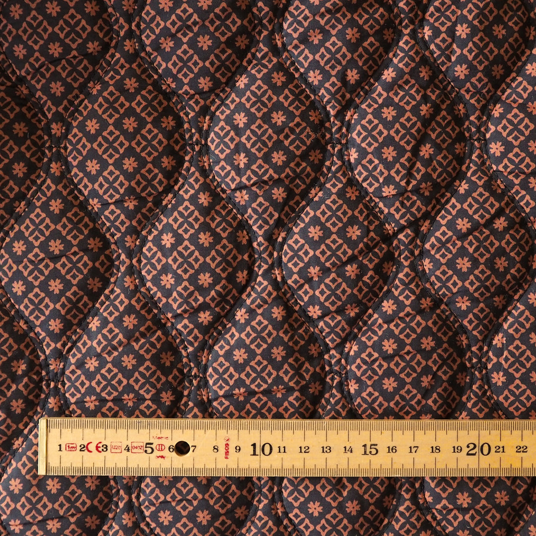 Close-up of a quilted fabric with a repeating geometric and floral pattern in dark navy, rust orange, and beige, with a wooden ruler at the bottom showing scale.
