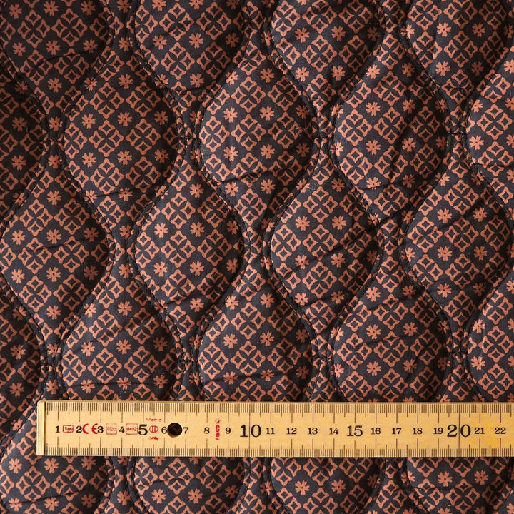 Close-up of a quilted fabric with a repeating geometric and floral pattern in dark navy, rust orange, and beige, with a wooden ruler at the bottom showing scale.
