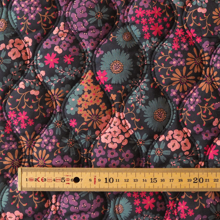 A close-up of a quilted fabric with a floral pattern in purple, pink, teal, and orange on a dark background, with a wooden ruler at the bottom showing the scale.