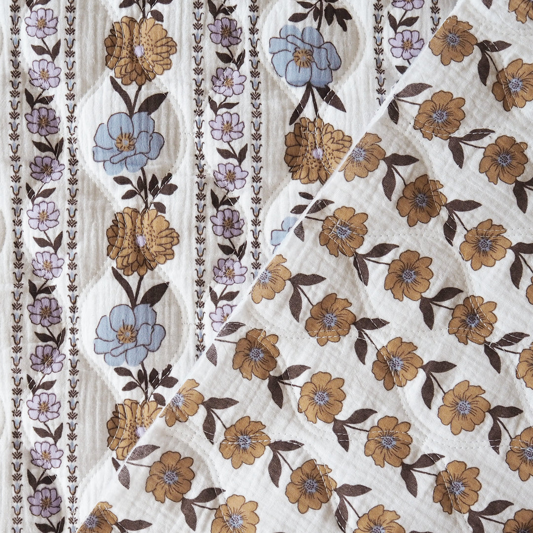 A close-up of a white muslin fabric with a delicate floral print featuring brown, tan, and light blue flowers with dark brown leaves, alongside vertical stripes of small purple and blue flowers.
