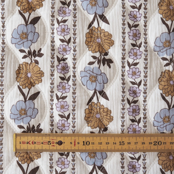 A close-up of a quilted fabric with a vertical floral pattern featuring blue, tan, and lavender flowers on a cream background, with a wooden ruler at the bottom indicating scale.