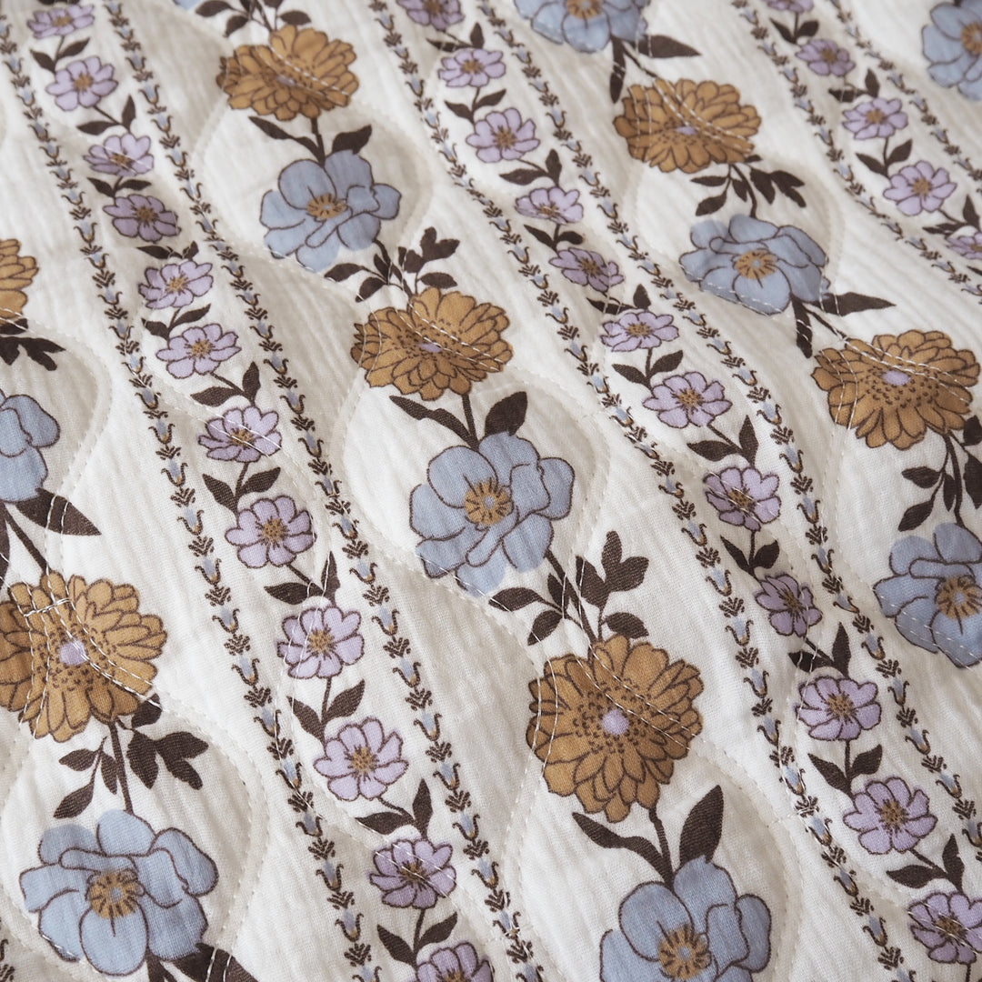 A close-up of a quilted fabric featuring a vertical striped pattern with large tan flowers, light blue flowers, and dark brown floral borders on an off-white background.