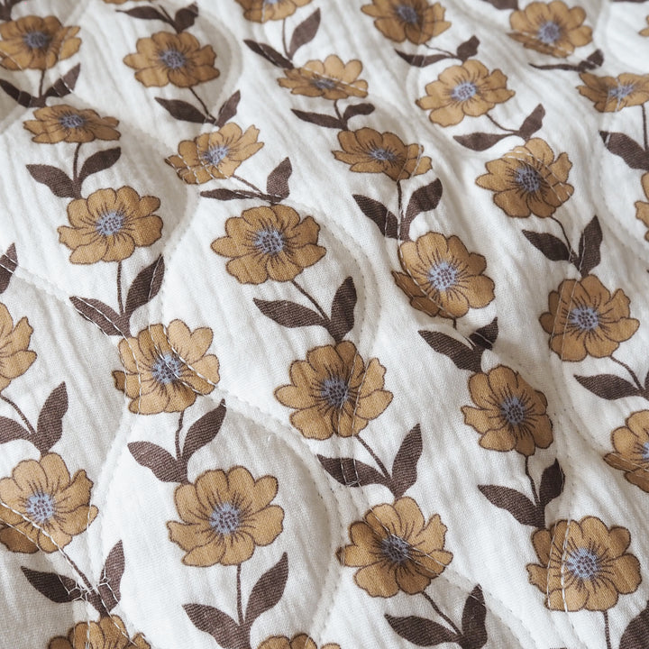 Close-up of a quilted white fabric featuring a repeating pattern of mustard yellow flowers with brown leaves and blue-purple centers.