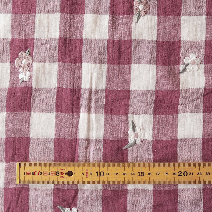 A close-up of a pink and white gingham fabric with small embroidered flowers, shown with a wooden ruler along the bottom edge for scale.