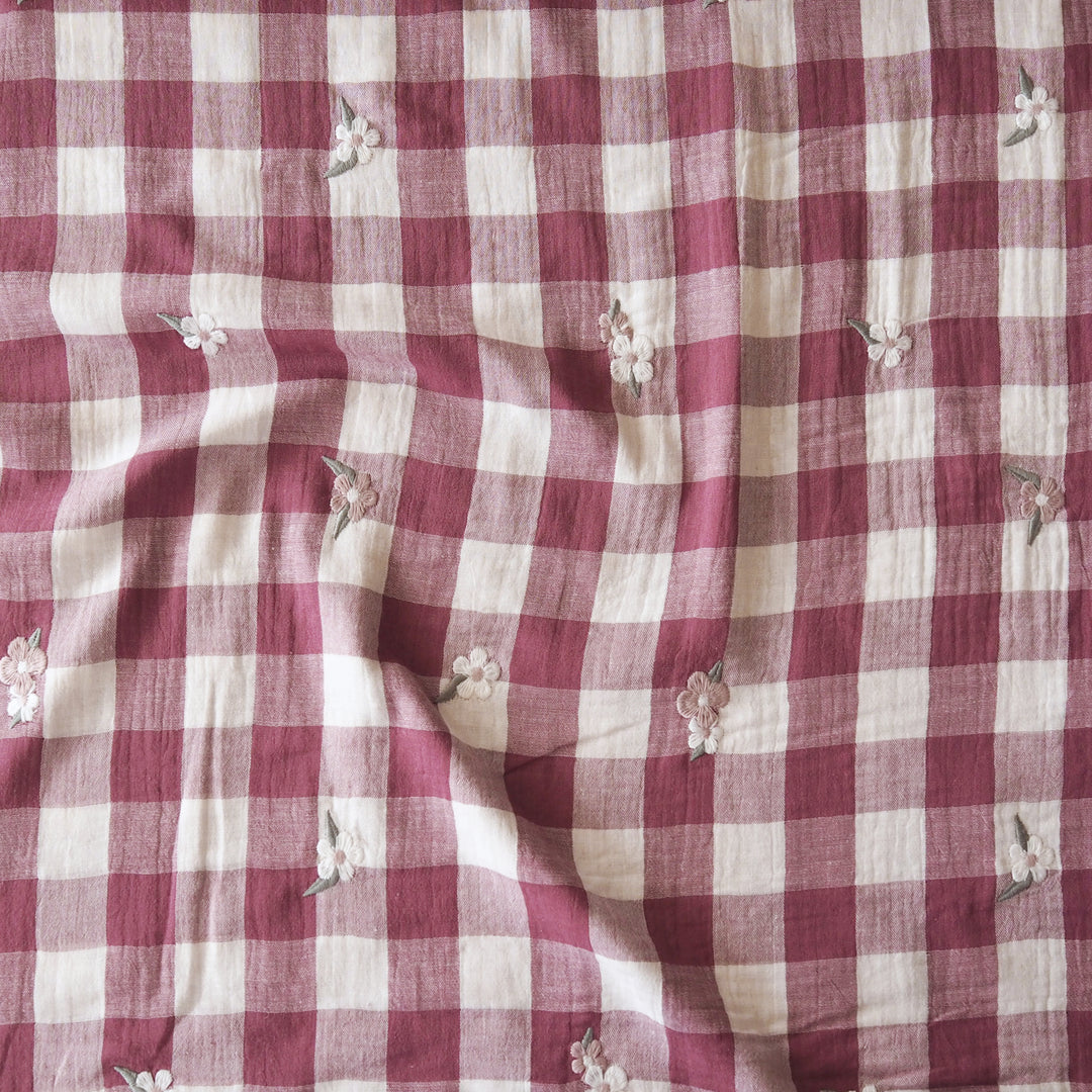 A close-up of a red and cream gingham fabric with small embroidered white flowers and green leaves scattered across it.