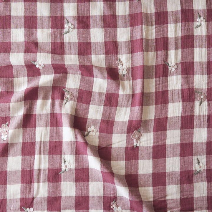 A close-up of a red and cream gingham fabric with small embroidered white flowers and green leaves scattered across it.